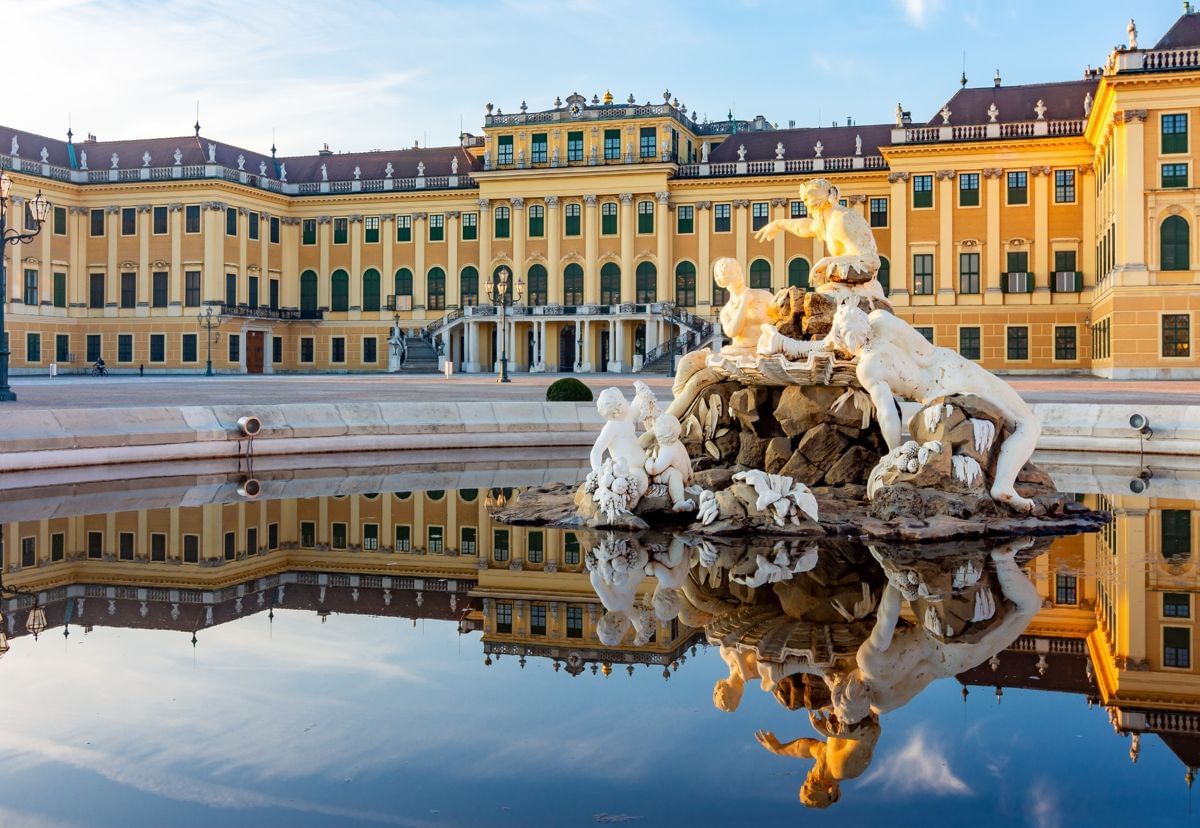 View of the grand yellow façade of Schönbrunn Palace in Vienna, with the ornate Neptune Fountain in the foreground and its statues.