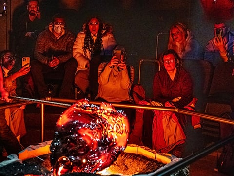 Audience members sit in close proximity watching a large, glowing, freshly-cooled lava formation, with several people photographing the mome