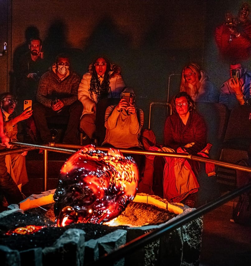 Audience members sit in close proximity watching a large, glowing, freshly-cooled lava formation, with several people photographing the mome