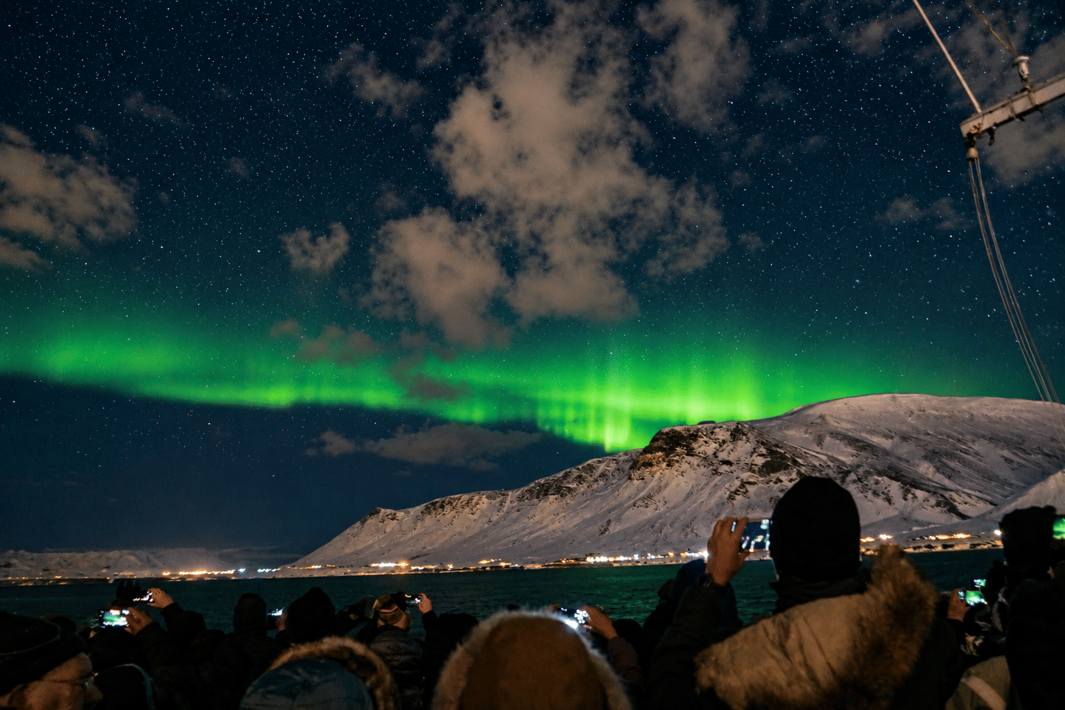 Passengers watching green Northern Lights beneath a cloudy night sky during a Reykjavík boat excursion.