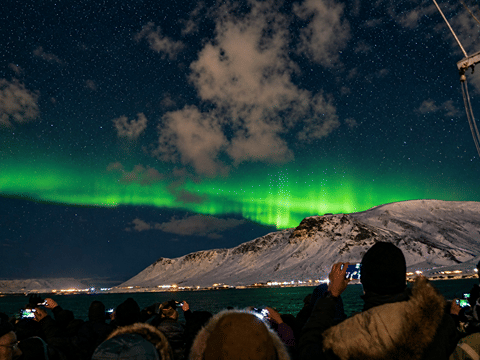 Passengers watching green Northern Lights beneath a cloudy night sky during a Reykjavík boat excursion.