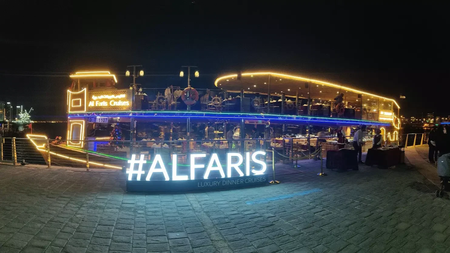 A large illuminated tour boat is docked next to a lit sign on a paved walkway at Dubai Water Canal Dhow Cruise.