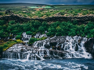 Dramatic dusk view of Hraunfossar waterfalls with deep green vegetation and dark lava rock, a small red-roofed farm visible on the highland