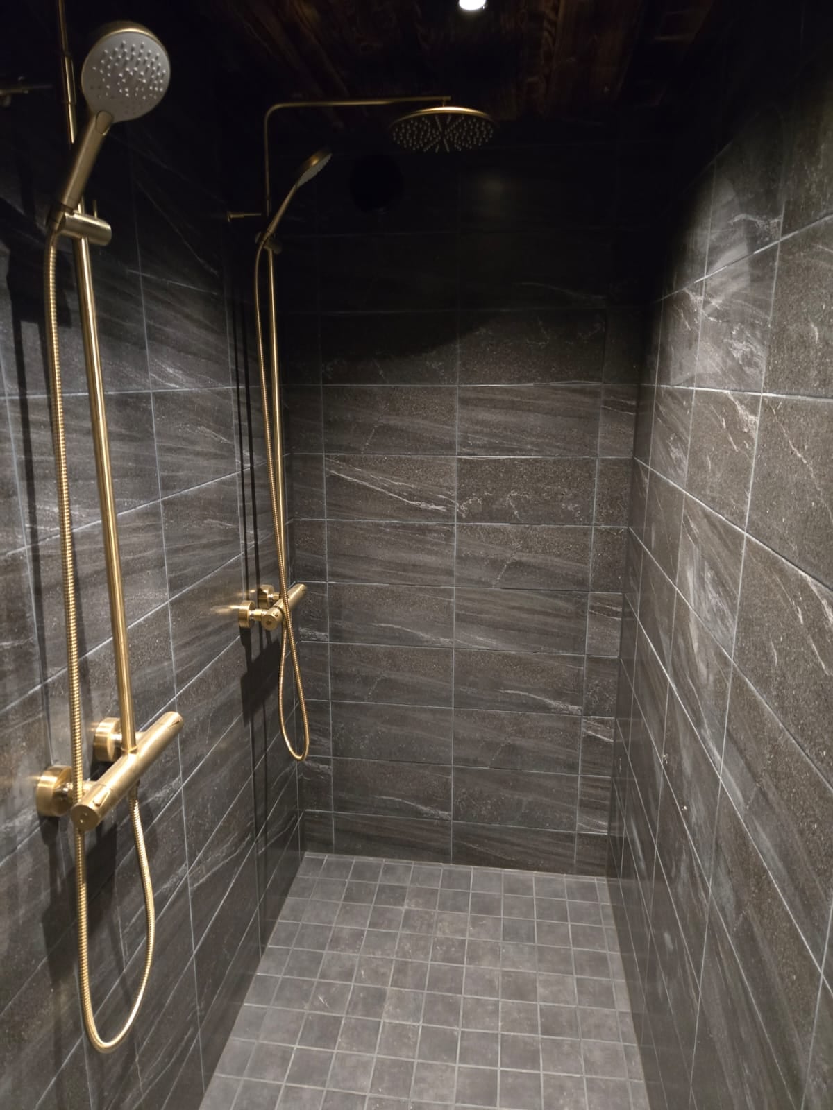 Private shower room with dark tiled walls and two brass shower fittings