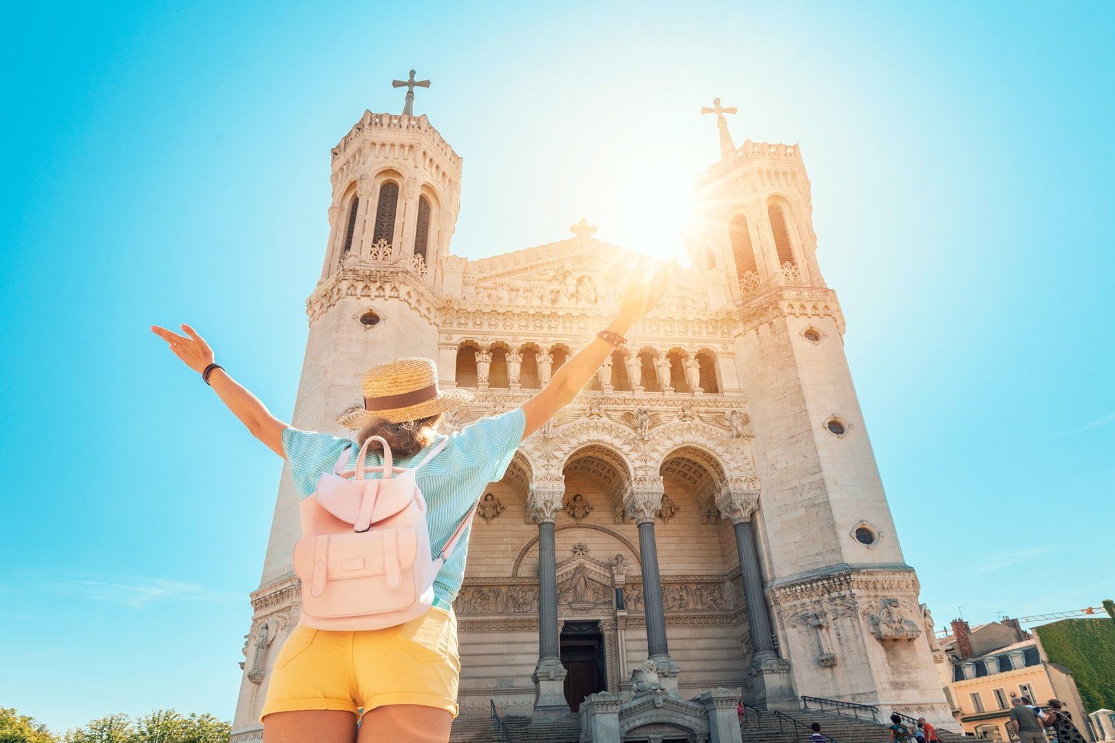 A joyful traveler with a backpack raises their arms in front of the Basilica of Notre-Dame de Fourvière in Lyon, with the sun shining above