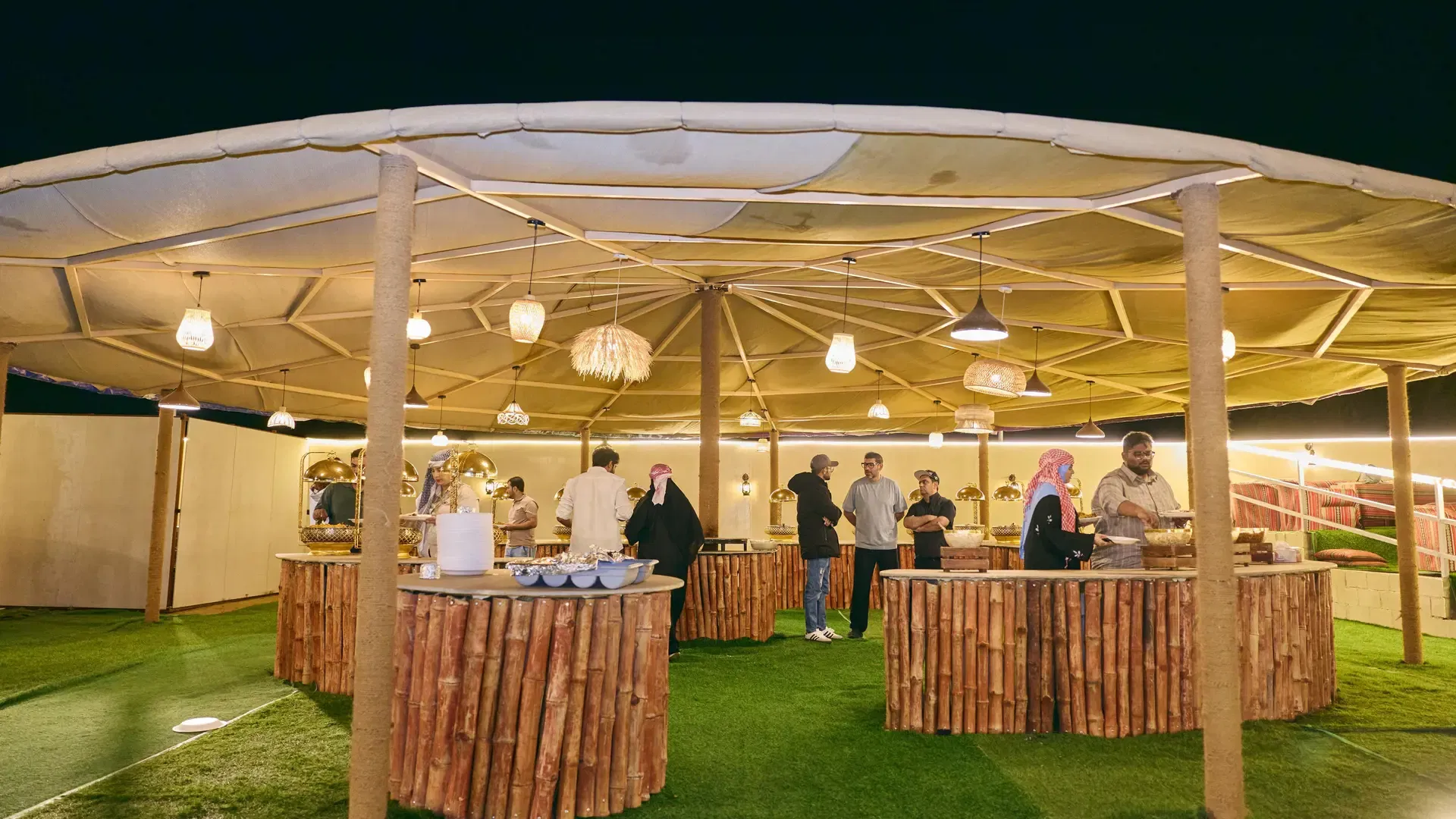 People gather around buffet counters under a large circular canopy draped with hanging lights at Desert Safari + Al Faqaa Camp.