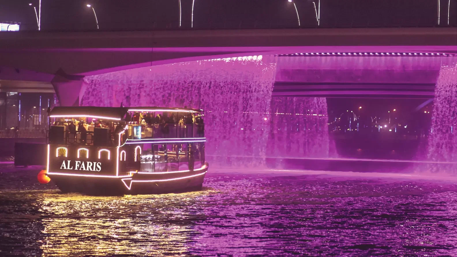 A double-decker cruise boat with glowing string lights navigates water near a waterfall lit in vibrant purple beneath a bridge at Dubai Water Canal Dhow Cruise.