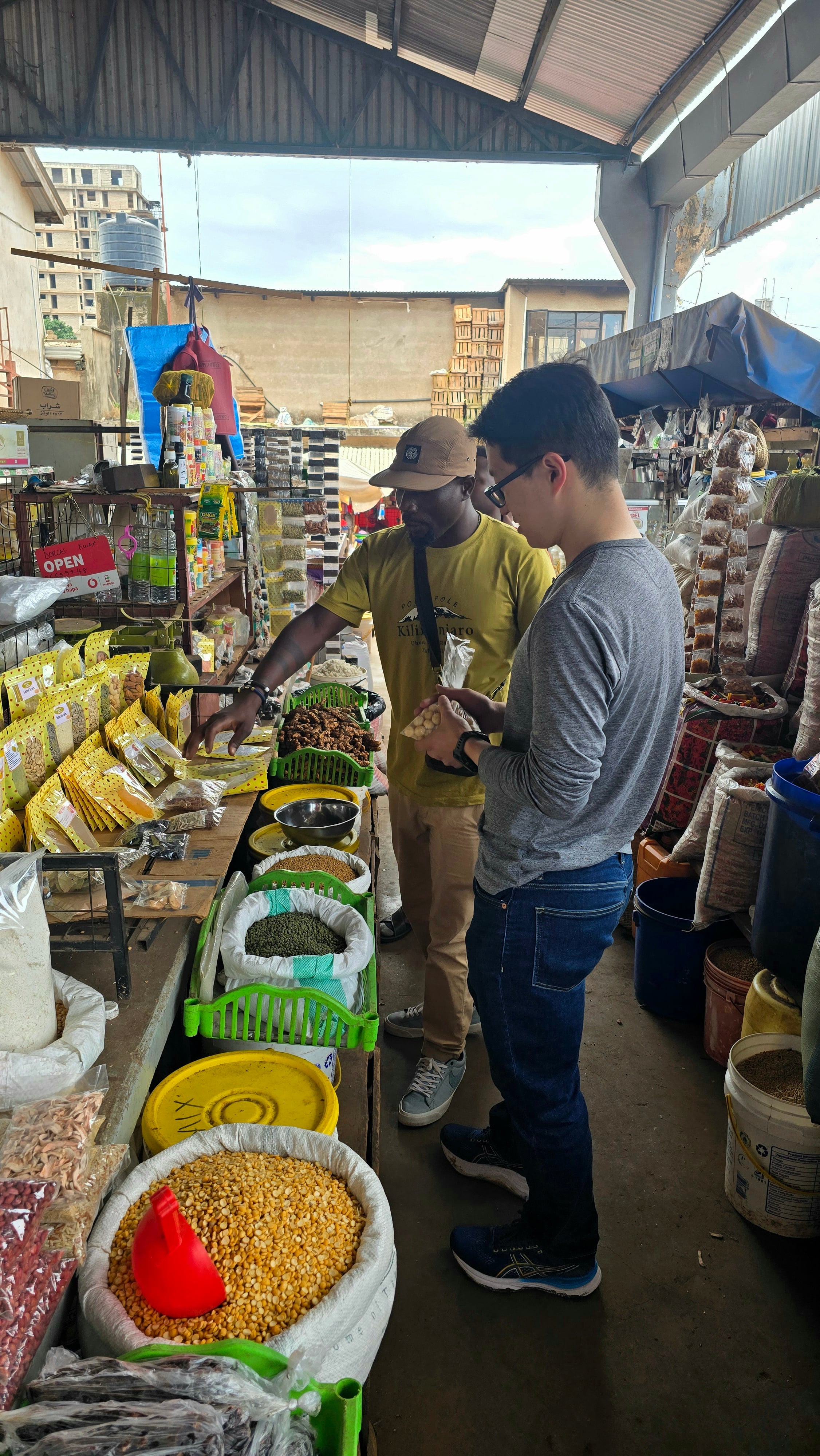 A guide assisting a traveler inside a covered market stall in Moshi filled with spices and local products.
