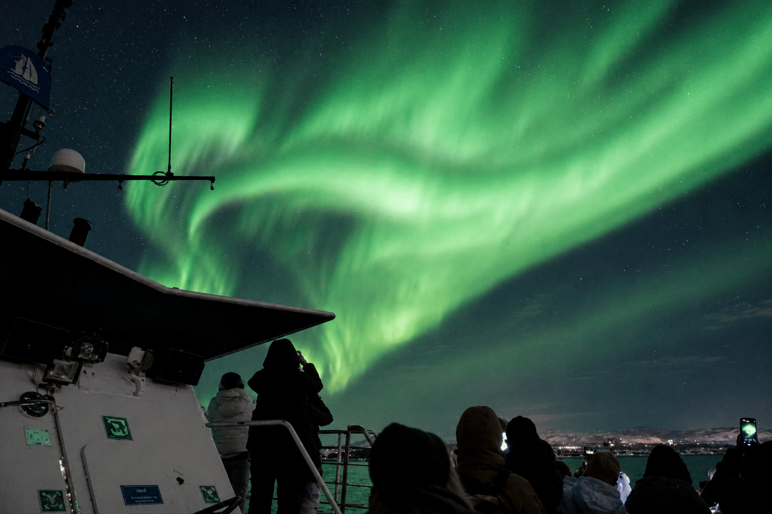 Passengers on a boat deck watching sweeping green aurora lights across the Icelandic night sky.
