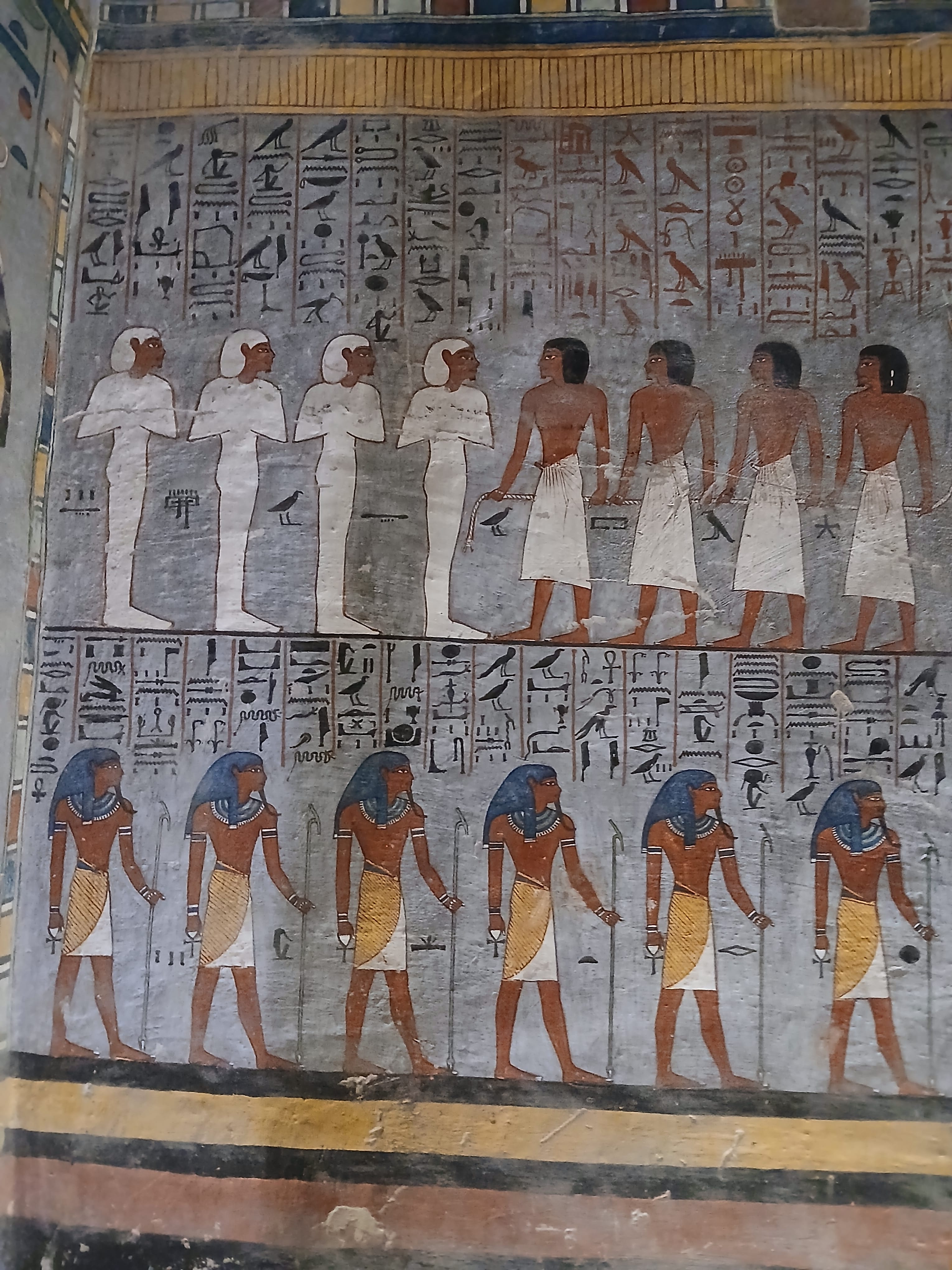 Two tiers of Ancient Egyptian figures and deities painted in blue, red, and white on a stone wall 