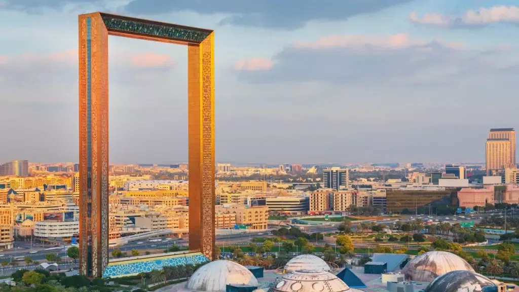 The golden rectangular tower of Dubai Frame rising above the city rooftops in warm evening light, with domed pavilions in the park below at Dubai Frame