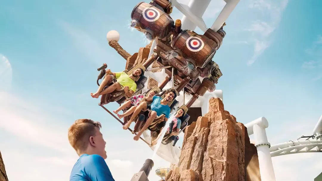Several people ride a suspended roller coaster past rocky scenery as a child watches from the foreground at Yas Waterworld, Abu Dhabi.