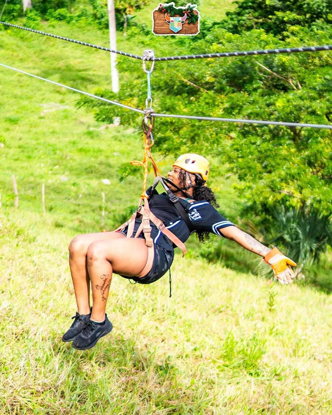 Woman wearing a helmet and safety harness gliding on a zip line over lush greenery in Punta Cana, extending her arms and enjoying the experi