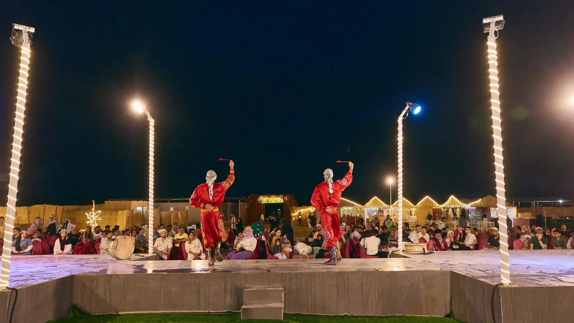 Two performers in red outfits dance on an illuminated stage before a seated crowd at Desert Safari + Al Faqaa Camp.