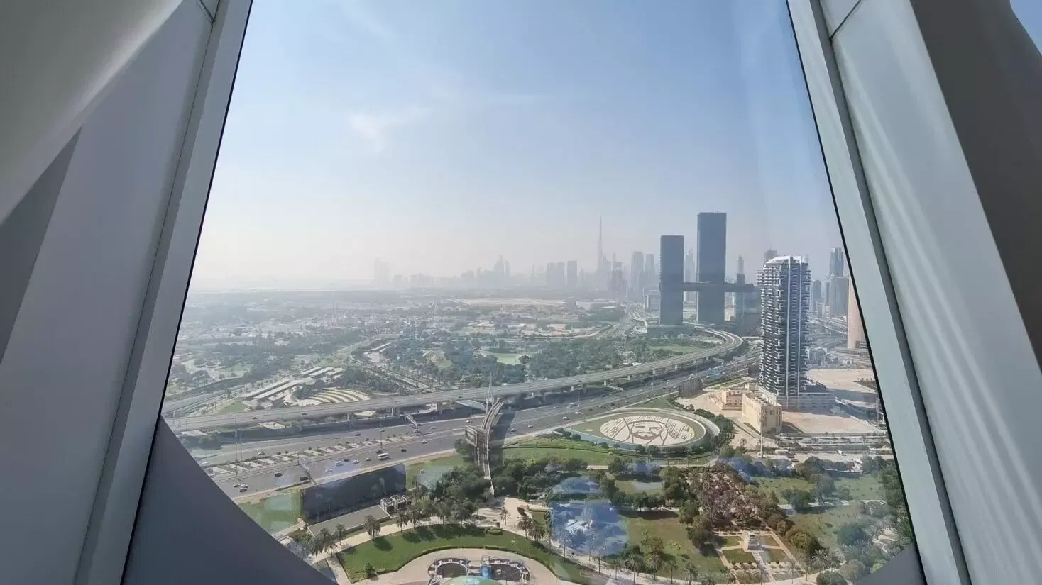 A hazy daytime view over parkland, motorways and the distant high-rise skyline seen through the angled deck windows at Dubai Frame