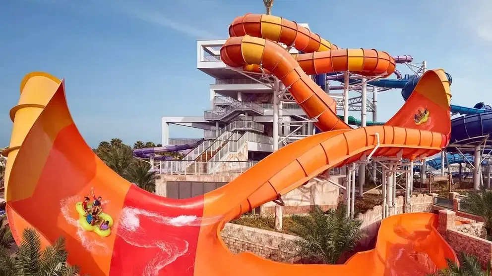 Bright orange and yellow slide flumes coiling around a white ride tower, with a raft of riders splashing through a wide funnel drop, at Aquaventure Waterpark