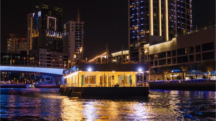 A brightly lit boat moves along a canal past towering buildings and a bridge at night at Alexandra Sea Lounge.
