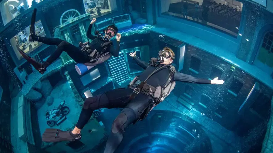 Two divers in wetsuits and scuba gear descend into a deep, flooded urban set featuring a submerged motorcycle and detailed building facades at Deep Dive Dubai.