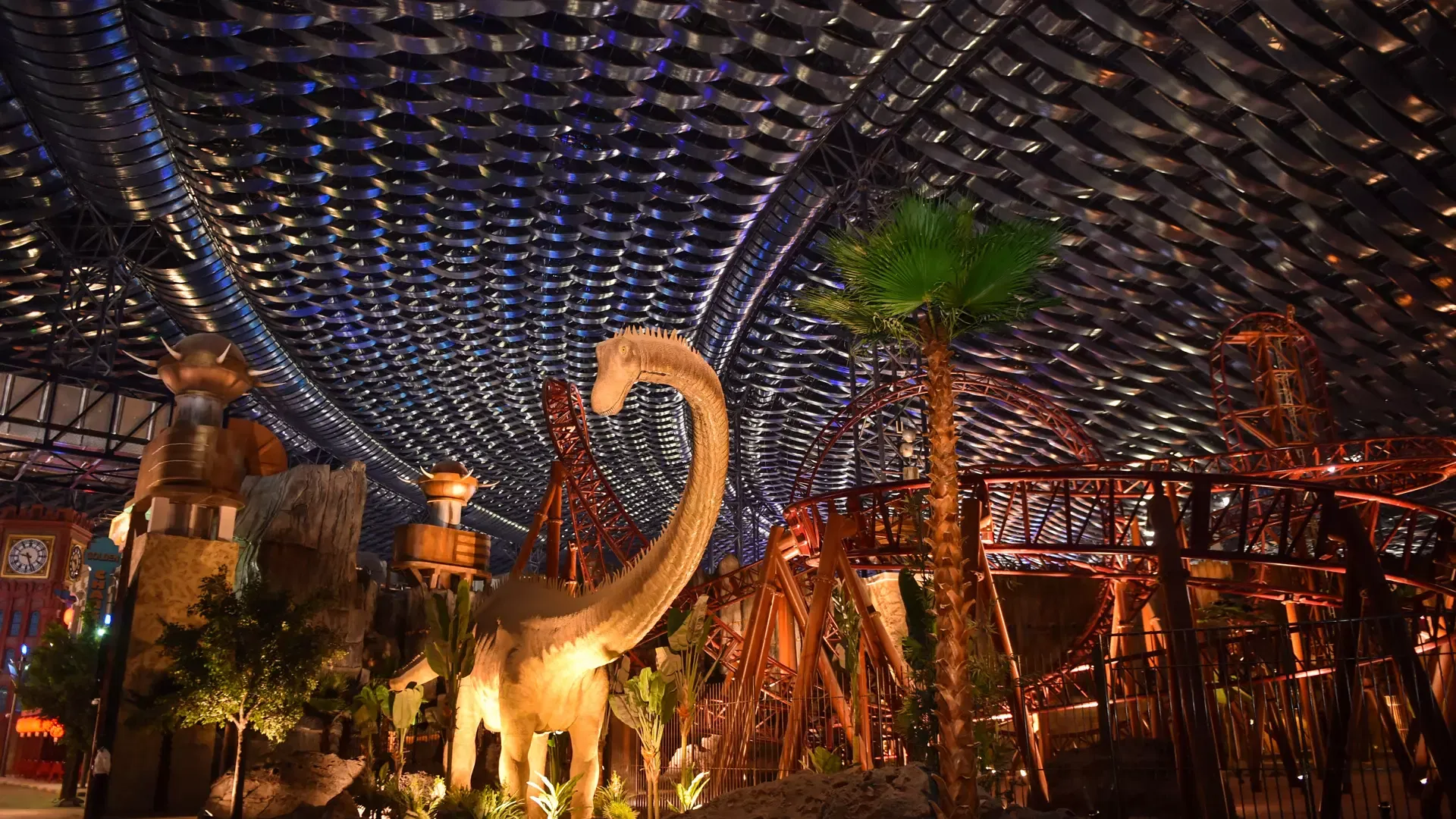 A large dinosaur figure stands inside a cavernous, dark hall beneath an undulating metal ceiling with roller coaster tracks winding overhead at IMG Worlds of Adventure Theme Park.