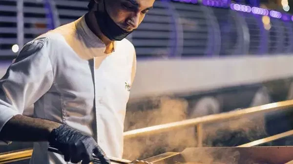 A chef wearing a white uniform, black mask, and gloves cooks over a steaming grill at Alexandra Sea Lounge.