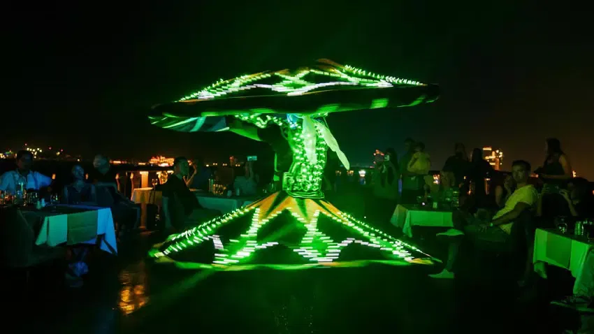 A performer in a spinning skirt illuminated by bright green lights entertains diners seated at outdoor tables at night at Alexandra Dhow Cruise in Dubai Marina.