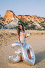 A woman in her thirties smiles broadly on a beach, holding a "30" balloon. A small cake sits on a wooden surface in the foreground against t