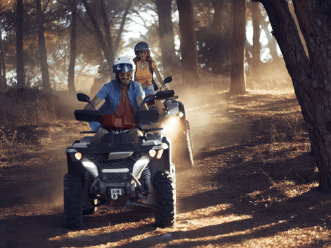 Two ATV riders in helmets navigating a dusty dirt track through sun-dappled trees, headlights on, riding in convoy.