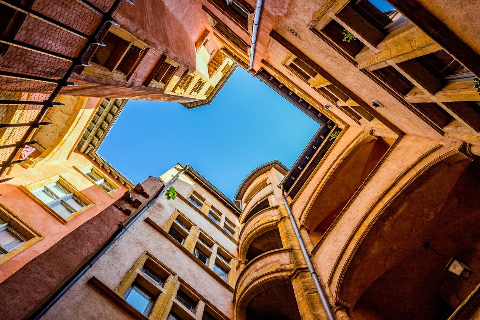 Upward view of a historic traboule in Lyon, showcasing warm-toned Renaissance buildings with intricate arches and a bright blue sky above.
