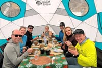 A group of six hikers smiling and enjoying a meal together around a set table inside a large white and teal geodesic dome tent.