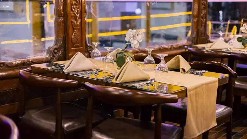A dining table with folded tan napkins, wine glasses, and a small vase of white flowers sits next to an ornately carved wooden partition and windows at Alexandra Dhow Cruise in Dubai Marina.