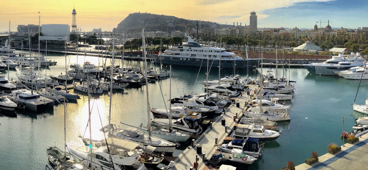 Standing in front of sailing boats at the harbour. In the back you will find the shape of Montjuic hill where the tour will take place.