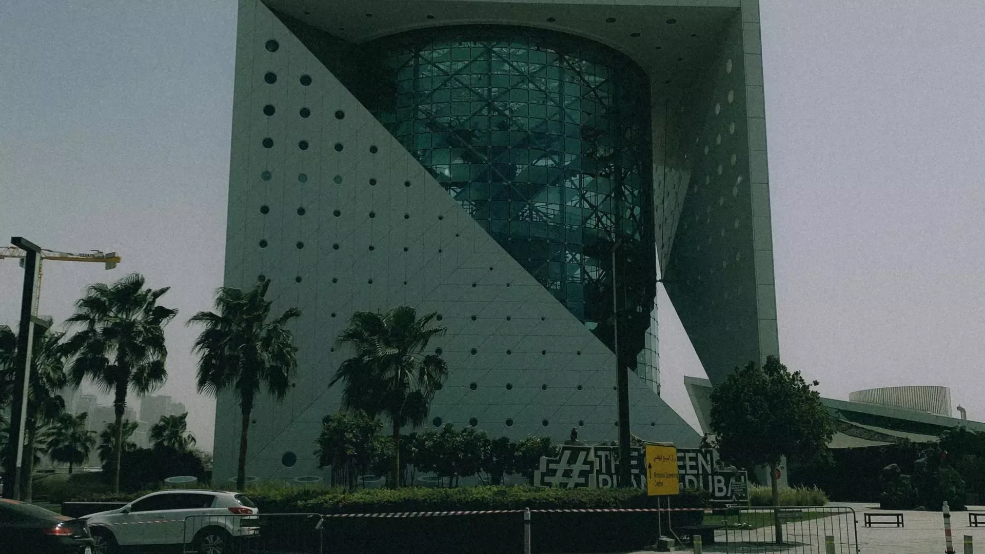 The exterior of The Green Planet Dubai, a cube-shaped building with a glass rainforest visible inside, framed by palm trees