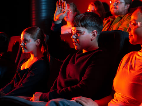 A young boy wearing safety glasses raises his hand while sitting in a dark theater alongside other children and adults, illuminated by the r