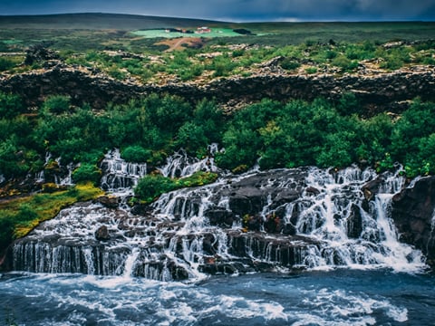 Dramatic dusk view of Hraunfossar waterfalls with deep green vegetation and dark lava rock, a small red-roofed farm visible on the highland