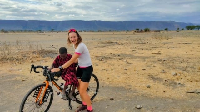 A female cyclist standing with her bicycle next to a young Maasai child in traditional red shúkà outdoors with hills in the background.