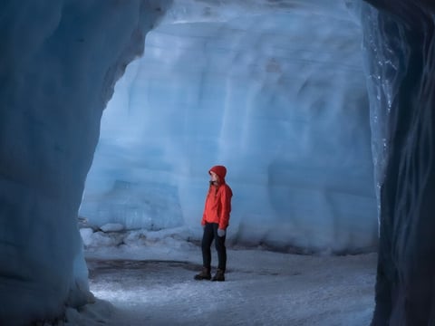 A lone visitor in a red jacket and hood standing inside a large chamber within the Langjokull glacier ice tunnel system, surrounded by tower