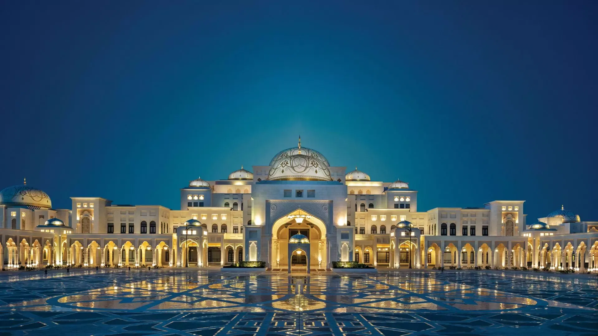 A wide shot of the grand palace exterior under a deep twilight sky, highlighting the intricate white and gold architectural details, glowing windows, and the reflective plaza surface at Qasr Al Watan, Royal Palace.
