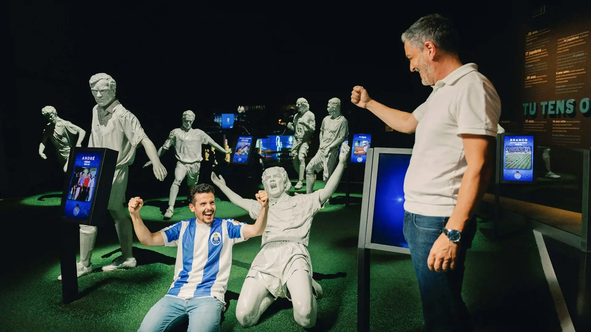 A smiling man sits on the floor next to statues in a dimly lit sports exhibition room at FC Porto Museum & Dragão Stadium ticket.