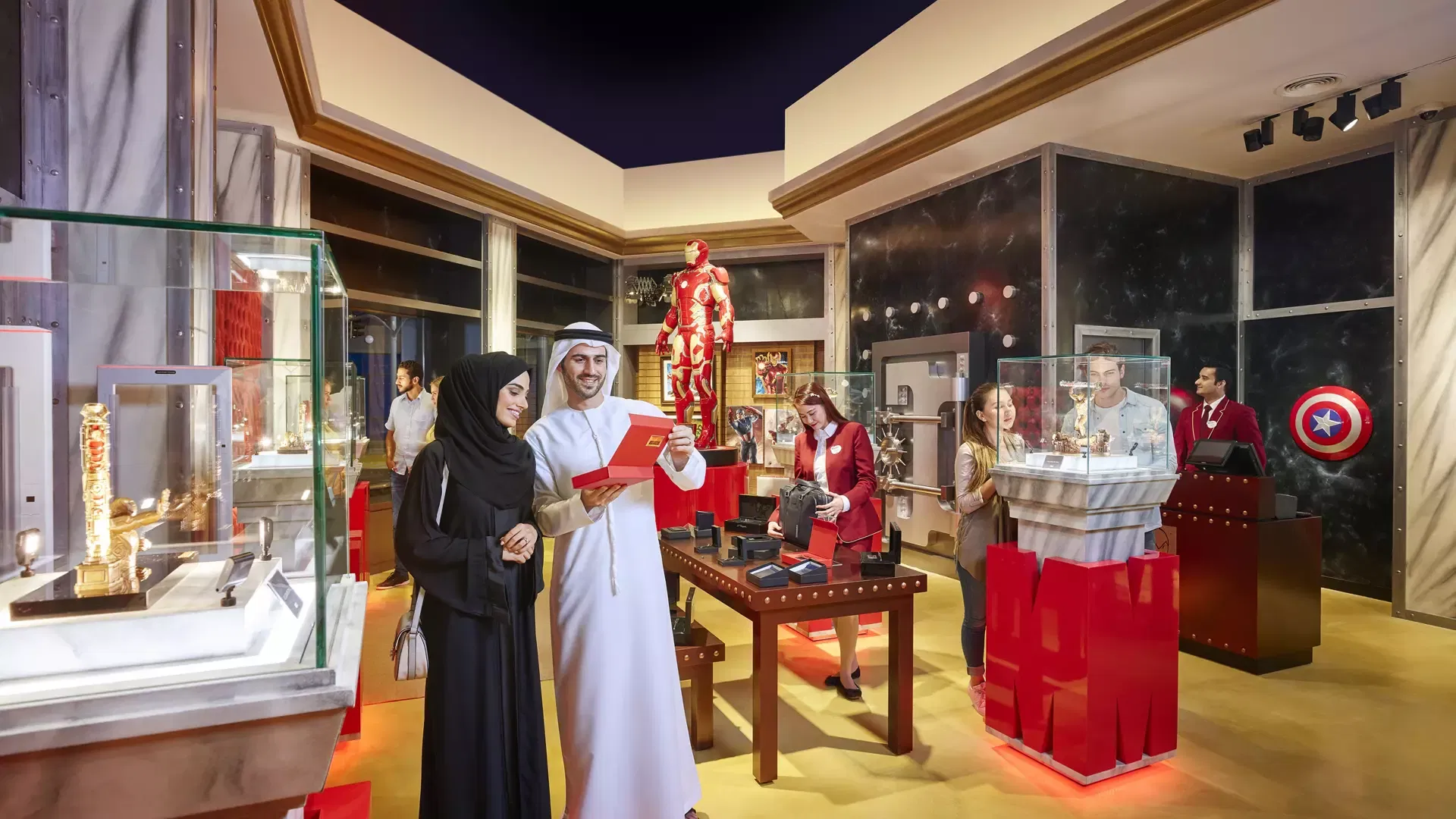 A man in traditional white attire and a woman in a black abaya smile while holding a red box in a brightly lit merchandise store filled with display cases, a red robotic suit, and visitors browsing at IMG Worlds of Adventure Theme Park.