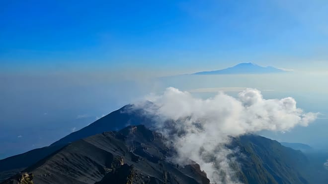 A sharp, dark volcanic mountain ridge with white clouds drifting over the slopes under a clear blue sky.