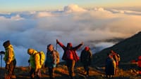A group of trekkers with backpacks standing on a mountain ridge above a thick sea of clouds at sunrise.