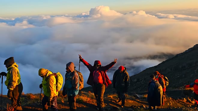 A group of trekkers with backpacks standing on a mountain ridge above a thick sea of clouds at sunrise.
