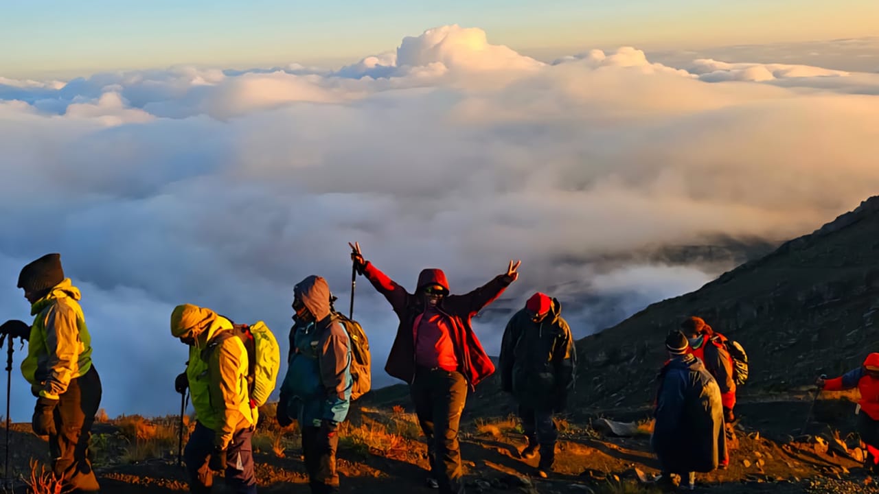 A group of trekkers with backpacks standing on a mountain ridge above a thick sea of clouds at sunrise.