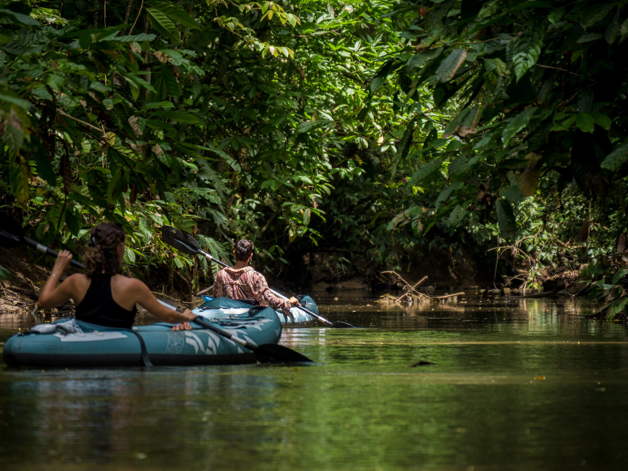 Two people paddling inflatable kayaks along a calm jungle river with dense green rainforest on both sides.