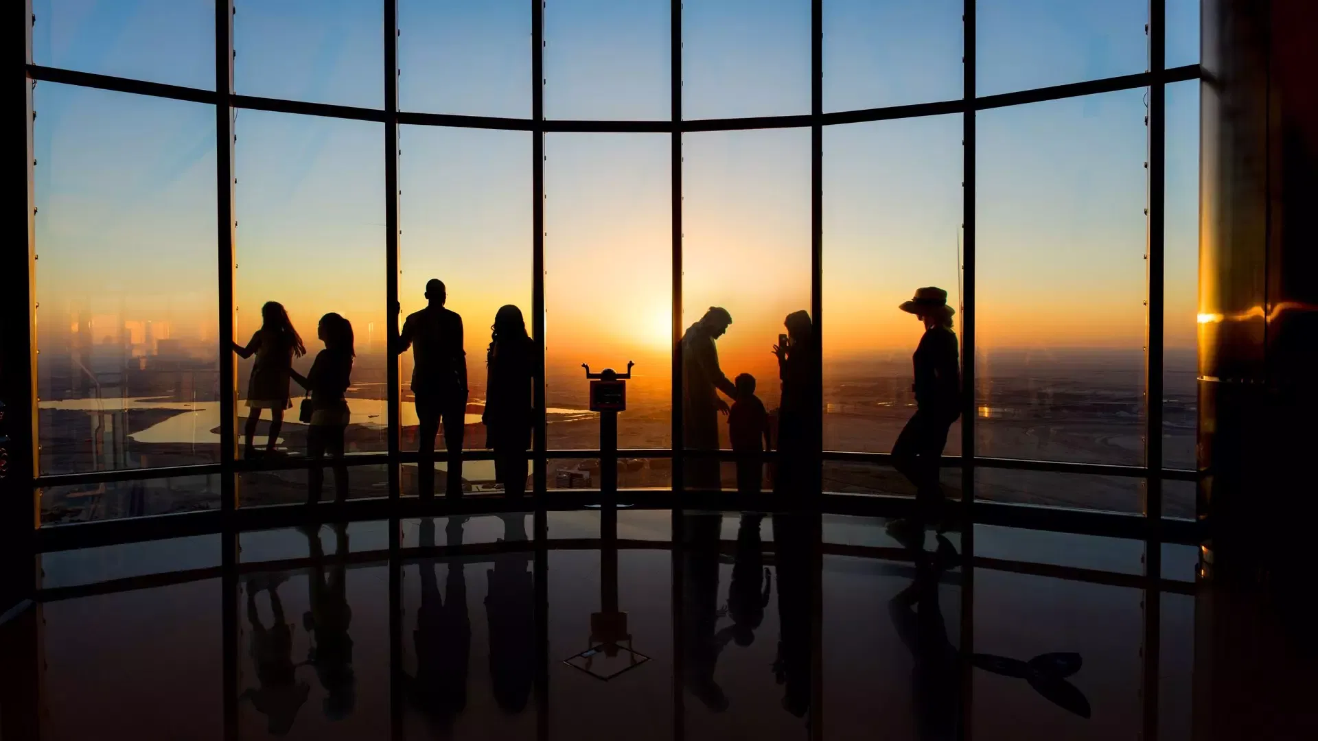 Families watching the sun drop to the horizon through the tall window bays of the observation deck, a viewing telescope standing between them, at Burj Khalifa