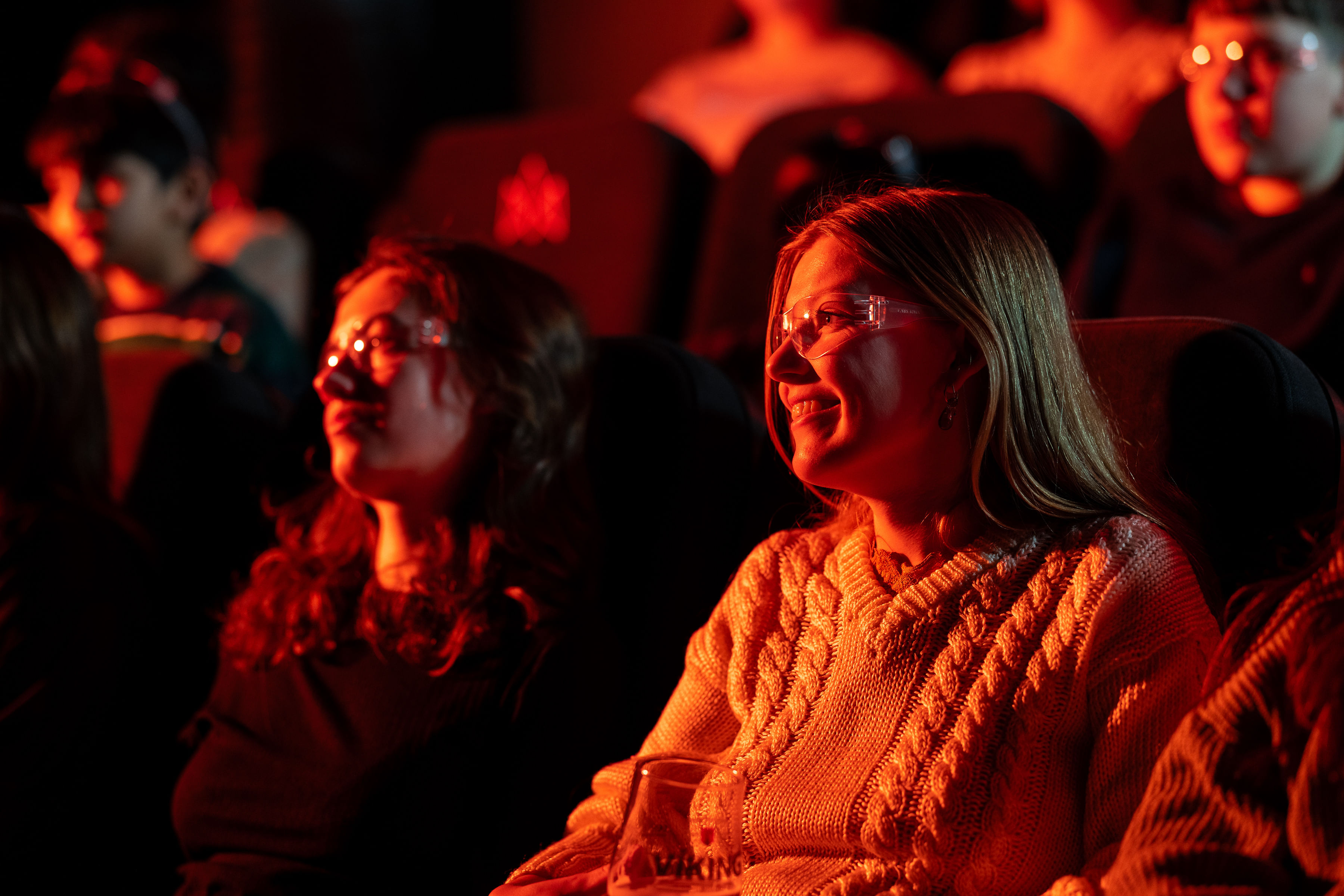 Two women wearing safety glasses smile as they watch the Lava Show, lit warmly by the red and orange glow from molten lava.