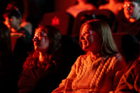 Two women wearing safety glasses smile as they watch the Lava Show, lit warmly by the red and orange glow from molten lava.