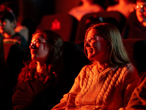 Two women wearing safety glasses smile as they watch the Lava Show, lit warmly by the red and orange glow from molten lava.