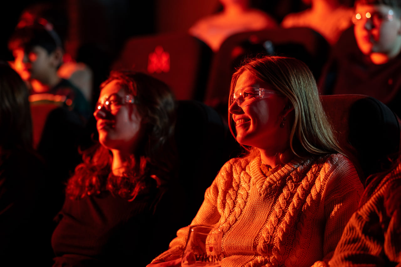 Two women wearing safety glasses smile as they watch the Lava Show, lit warmly by the red and orange glow from molten lava.