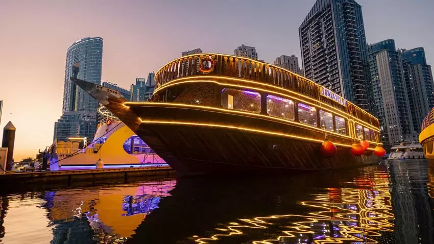A wooden dhow ship outlined in glowing yellow lights is docked along a pier at dusk at Alexandra Dhow Cruise in Dubai Marina.